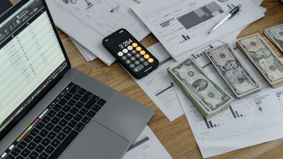 Стос доларових купюр поруч із калькулятором на смартфоні та ноутбуком з таблицею