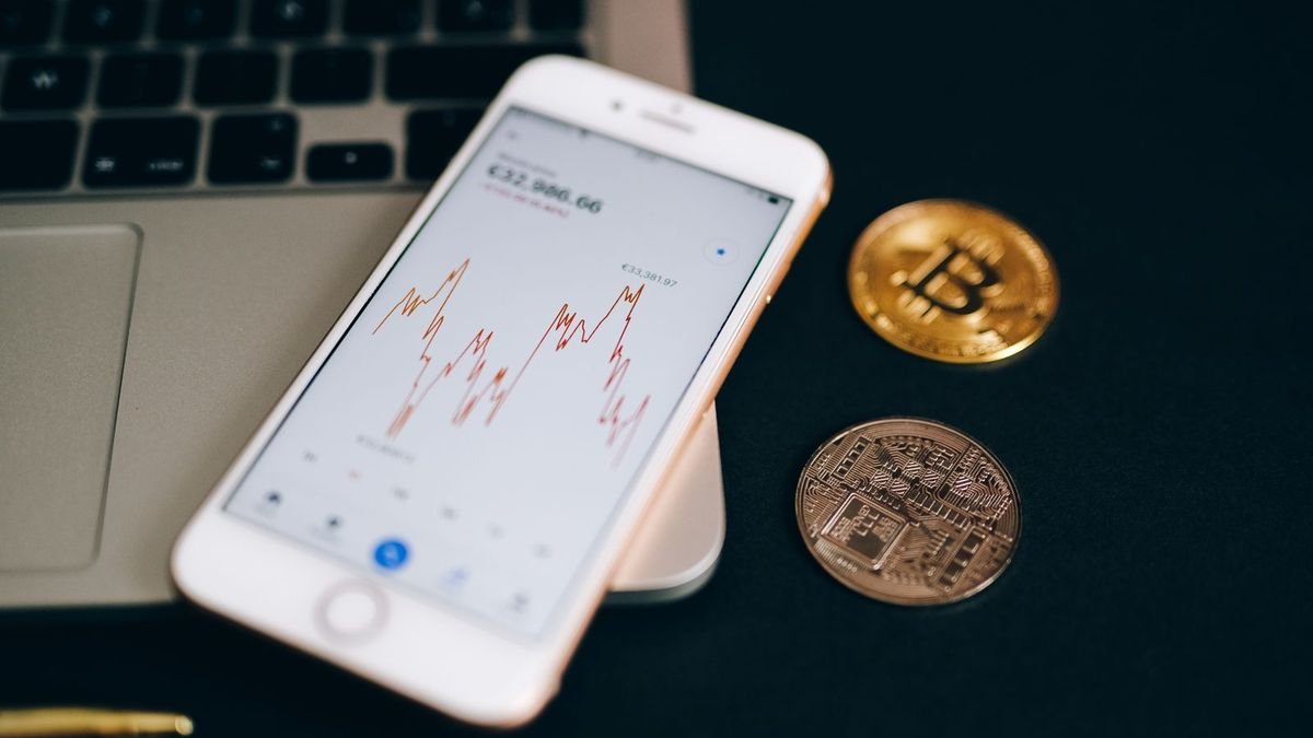 Смартфон із графіком на екрані та дві монети біткоїна на чорній поверхні біля ноутбука